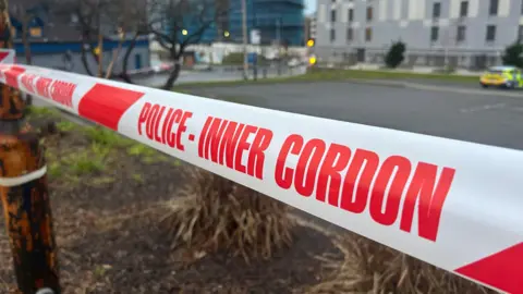 Red police cordon tape in the foreground and a police car in the background. There are buildings in the background as well.