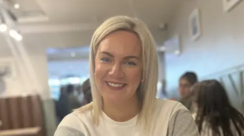 Sarah Harrigan Sarah Harrigan in a restaurant with people in the background. She has blonde hair and is wearing a white and grey sweater. 