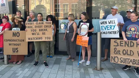 BBC A line of parents outside a council office building hold placards saying with slogans including: "When will every child matter?"