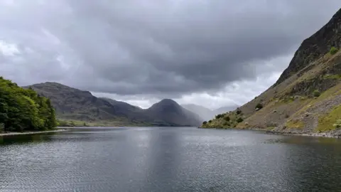 posiden A photo which appears to be either taken next to or actually on a lake. Grey water spreads out in front and disappears around the corner of a mountain which is jutting out into the shot. In the distance, more mountains become obscured behind fog. Clouds above are growing heavier and darker.