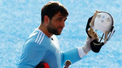 George Pinner with one arm lifted up and holding up a helmet and a hockey stick under his other arm. He is wearing a long-sleeved and light blue Adidas sports shirt. He has dark hair and a short beard.
