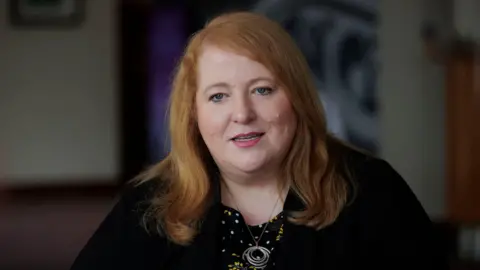 PA Media Naomi Long looking at the camera. She is wearing a black top and a silver necklace. The background is out of focus.