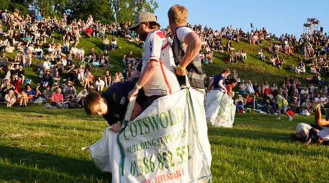 PA Three young men standing in a large industrial plastic bag which says 'Cotswold Building Supplies' on the side. They are competing in a sack race during the Cotswold Olimpick Games. There are large crowds of spectators in the background, sitting on a grassy hill. It appears to be approaching dusk and the light is low and slightly orange.