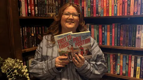 A woman with long auburn hair is smiling and holding two books. She is standing in front of bookshelves filled with colourful books. She is wearing a light blue shirt with dark blur stripes. The books in her hands are titled "first to finish" and "pole position"