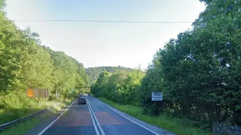 The A590 with two cars in the left lane and a sign on the right reading 'Newby Bridge Please drive carefully' 