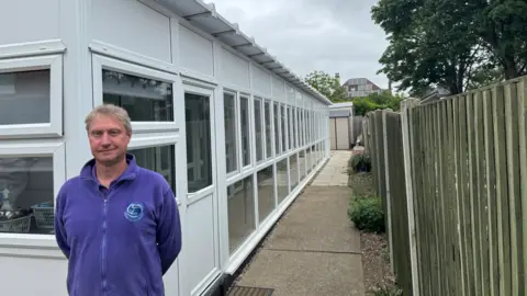 George King/BBC Nick Chaplin standing in front of the rescue centre's state-of-the pens He is wearing a purple jumper with the charity's logo on it.