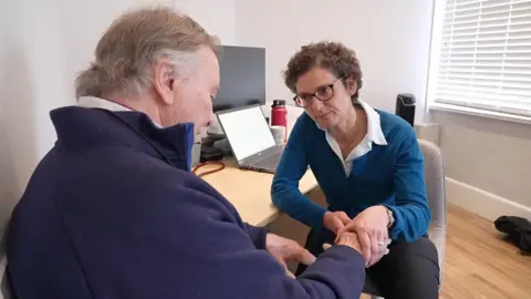 BBC/MARTIN GILES Patient Ian Miller is seen by Dr Yvonne Girgis‑Hanna in her clinic room. It is a small modern room with a water machine in the background. There is light wood laminate flooring and a white window blind. She is sitting on a modern beige chair and has a laptop on the desk with a traditional computer and a water bottle in the background. She is examining Ian's hand. Ian has arthritis in the hand he draws with. She is wearing an ink blue jumper and white blouse and has short brown curly hair and glasses and he has grey hair and a navy fleece on.