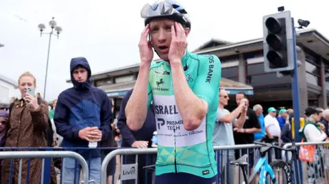 A man wearing a cycle helmet and a turquoise cycling outfit holds his head in his hands as he wins the Tour of Britain. Crowds behind him and barriers line the street