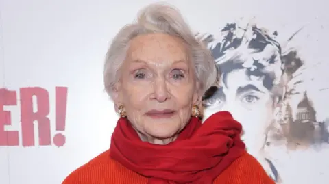 Yui Mok/PA Media An elderly woman, Sian Phillips, who has grey hair, is wearing a red scarf and an orange top.