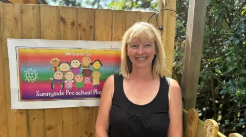 A lady in a black vest top stands in front of a wooden fence with the sign to a preschool attached to it. 