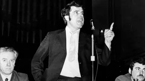 Getty Images A black-and-white photo of a Jimmy Reid, who has dark hair and side burns, speaking into a microphone. He is wearing a dark suit with a white shirt and patterned tie. He has his left hand raised in front of him, with his index finger pointing upwards. 