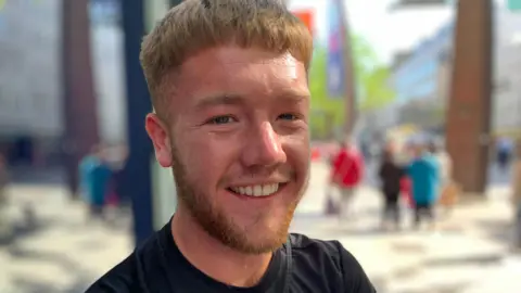 A man with short brown hair and a neat beard is smiling at the camera. He is wearing a black t-shirt.