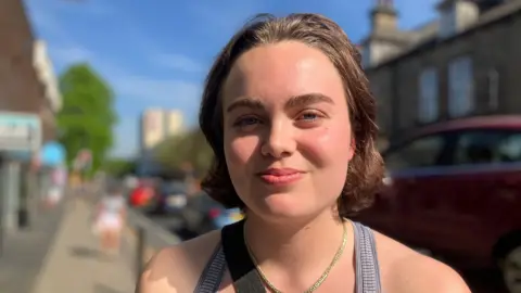 BBC/Elizabeth Baines A woman with short brown hair smiles. She is wearing a grey strappy top and a necklace. Behind her a row of shops is blurred.