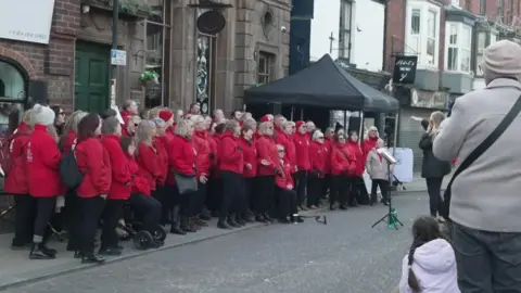 The choir wear red fleeces and black trousers.  The conductor stands at the front with a music stand.  Spectators stand in the foreground of the shot.