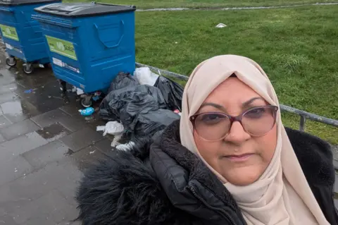 Sadia Khan Sadia Khan is wearing glasses and a light beige head covering. She has on a thick black coat and is standing next to overflowing bins in her local park. 