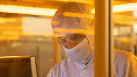 A scientists whose face and head is covered with protective clothing analyses something on a screen.