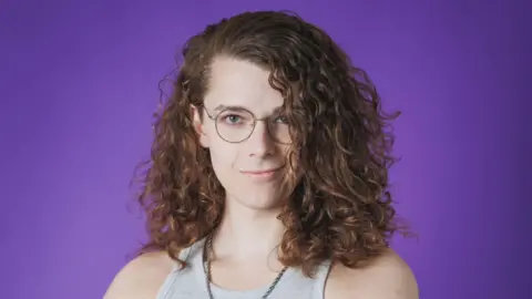 Alastair/Eret Eret, a streamer who has curly brown hair and glasses is staring at the camera, wearing a grey top. There is a purple background.