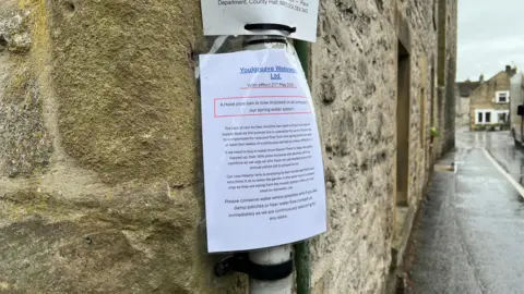 Image shows a sign put up in the village of Youlgreave in Derbyshire, reading: "The lack of rain for two months has seen a drop in our reserve supply level as the pumps are in operation for up to 5 hours a day to compensate for reduced flow from the spring which will need at least two weeks of continuous rainfall to make a difference.
If we need to buy in water from Severn Trent to keep the system topped up, their 38% price increase will severely dent our cashflow so we urge all who have not yet started to pay their annual yellow bill to please do so.
Our new Header tank is emptying by 8am so we ask that those who think it is ok to water the garden in the early hours to please stop as they are taking from the header system when at most need for domestic use.
Please conserve water where possible and if you see damp patches or hear water flow contact us immediately as we are continuously searching for any leaks."