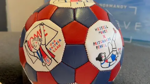 A football decorated by Pritchard with illustration of several Lionesses. One of the illustration is accompanied by an inscription reading "Hessell Makes x Pritchards Pencil".