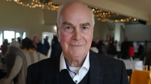 Qays Najm/BBC Mark Packnard standing inside a hall at a meeting. He is looking directly at the camera and smiling and is wearing a white shirt with a black jumper over the top and black suit jacket.