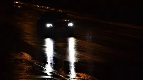 A car driving at night on a wet road, with bright white headlights reflected on the damp tarmac.