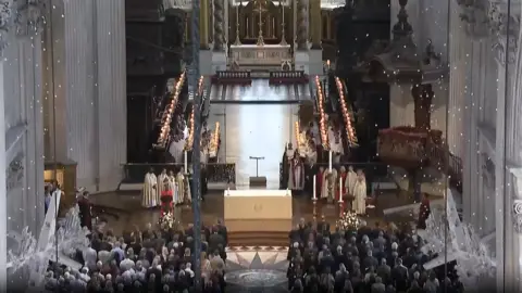 A wide shot of the inside of St Paul's Cathedral from above, with a full congregation and choir in place, as white petals fall from above.