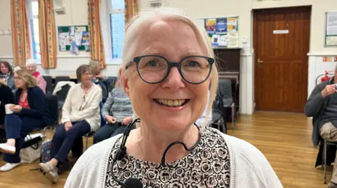 John Devine/BBC A woman standing in the middle of a room with a wooden floor and several chairs with people sitting on them. She has white hair in a ponytail, and is wearing green-framed glasses, a patterned top and a white cardigan.