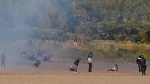 PA Media A disparate crowd of people stand in a cloud of gas, looking dejected, face Frenc police officers wearing riot gear near bushes along the treeline of a beach in Gravelines, France
