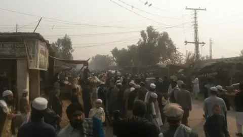 Maiwand Banayee یک خیابان شلوغ و گرد و غبار ، پر از مردان ، که بیشتر آنها لباس سنتی افغان را با کلاه های بی سیم پوشیده اند. ساختمانهای Ramshackle وجود دارد و پیلون ها خطوط برق را در پشت زمینه یک آسمان خاکستری نگه می دارند.
