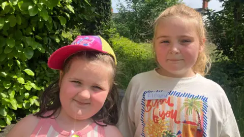 Two children standing together. One is short with dark hair and is wearing a cap. The other is slightly taller with blond hair in a ponytail. 