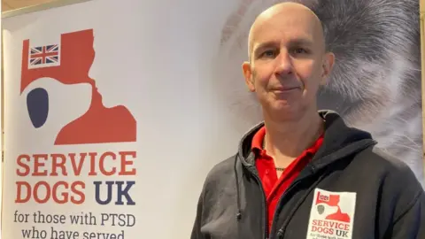 Nigel Rousell, Service Dogs UK, in front of a poster for the charity