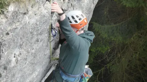 Aimee O'Doherty A climber scaling a rockface. They are wearing a white and orange helmet and a green top. There is a rope dangling from above. Alongside the rockface is a tree.