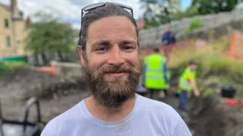 Lead arceologist, Dr Colin Elder, pictured on the dig site. He looks like he is in his 30s, has dark hair and a beard and has his glasses on top of his head. Behind him and out of focus are people in high-visibility jackets, taking part in the dig. 