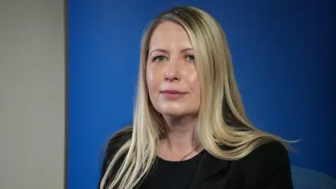 Leanne pictured looking into the camera. She's wearing a black blazer with a black top on underneath. She's sat in front of a royal blue backdrop with a white wall visible beyond it.