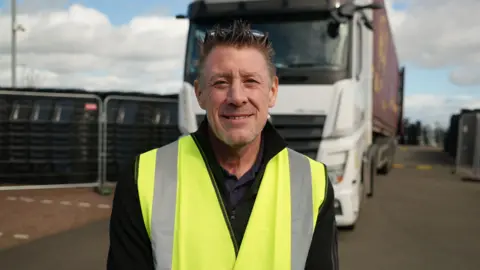 Qays Najm/BBC Head and shoulders image of John Exley looking into camera in front of a lorry and waste bins. He has spiky brown hair, a yellow hi-vis vest and a black jumper underneath.