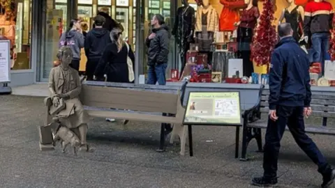 Image of the statue and bench superimposed onto an image of the pedestrianised shopping street. There are people walking along the street and in the background is the shop window of Goulds department store.