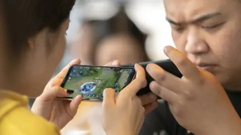 Getty Images Two people sit across from each other while playing games on there phones