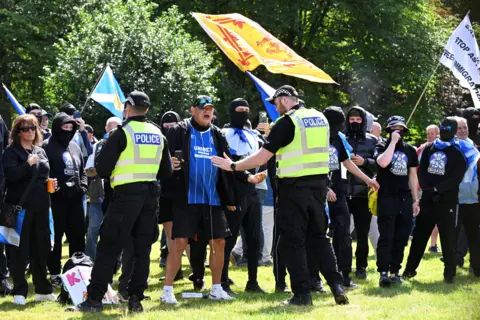 PA Media Officers attempt to calm a man during the anti-immigration demonstration, as a number of masked individuals wave flags in the background 