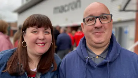 Leanne and Johnpaul attending the memorial service, she has dark hair and a fringe and is smiling into the camera, and on her right is her partner, who is bald, in a blue hoodie with dark rim glasses on.