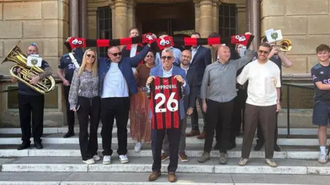 A group of people stand on the steps of the BCP Council Civic Centre. Some are raising red and black football scarfs. A man at the front is holding a Bournemouth strip. It has the number 28 on the back and the word "APPROVAL".