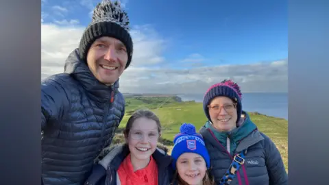 Mike Green Mike and Collette take a selfie with their two daughters during a walk along the coastline. They are all wearing winter clothing. It is a sunny day with some clouds in the background. The sea can be seen behind them. 