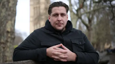 A man looks straight at the camera. He is wearing a black coat. The framing and content of the picture look sombre. 