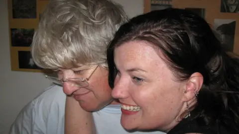 A young woman with brown hair embraces an older woman with glasses and blonde hair, in a hug together