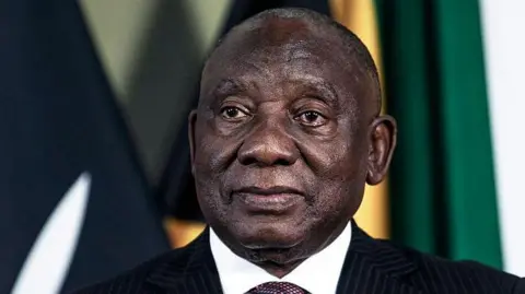 A head-and-shoulders image of Cyril Ramaphosa. He is wearing a dark suit and the white collar of his shirt can just be seen. Some flags are out of focus in the background.