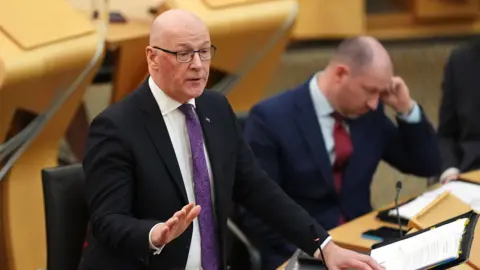 PA Media A bald man with glasses wearing a dark suit and a purple tie