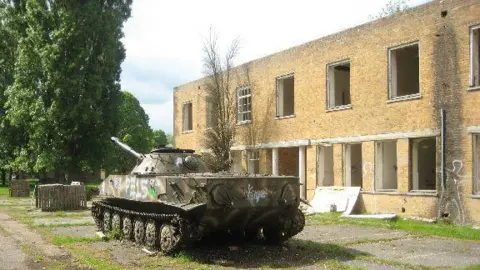 Peter Neal/Geograph Two-storey light brown brick building with square holes where the windows should be. There is a drain pipe to the right with a damp patch round it. A military tank is in the foreground, with graffiti on the side. Large fir trees can be seen to the left. The concrete base in front of the building has grass growing through it.