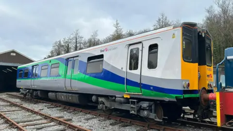 ACMU Society A single electric train carriage on a railway track in front of a storage building. It has been painted in grey, green and blue with some white as well as yellow on its front. 