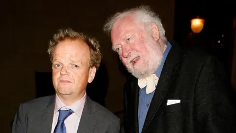 Toby Jones, in a grey suit pink shirt and blue tie, stood next to his dad. Freddie Jones is wearing a black suit, blue shirt and pink bow tie.