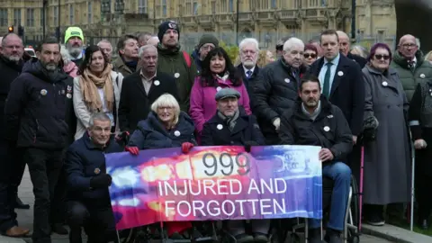 People stand behind a banner reading "999 INJURED AND FORGOTTEN"