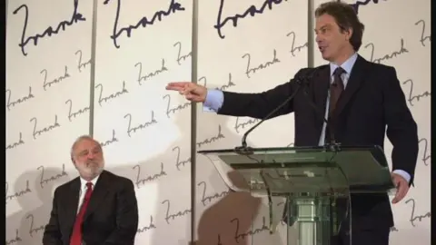 PA Media Tony Blair in a dark suit and brown tie stands at a podium with a 'Frank' themed background. Frank Dobson sits to his right wearing a dark suit and red tie.